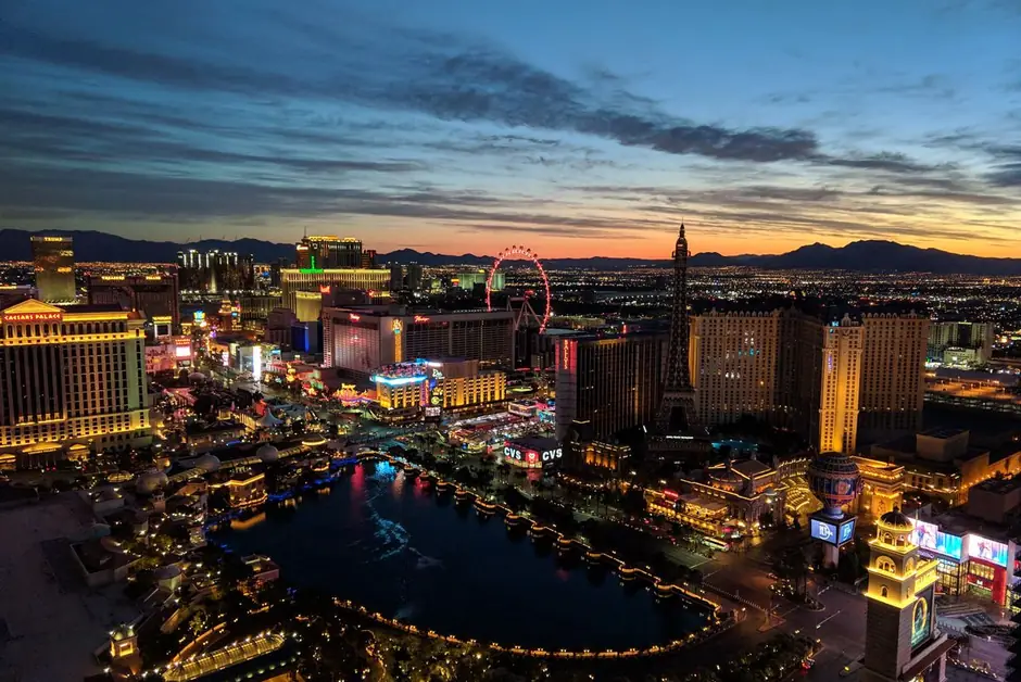 The Las Vegas Strip at dusk, the Bellagio fountains lake and the High Roller wheel under a blue and orange sky.