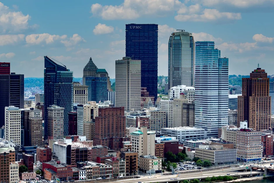 Downtown Pittsburgh from the air: the dark UPMC tower and the stepped BNY Mellon and PPG buildings above the brick blocks of the Golden Triangle.